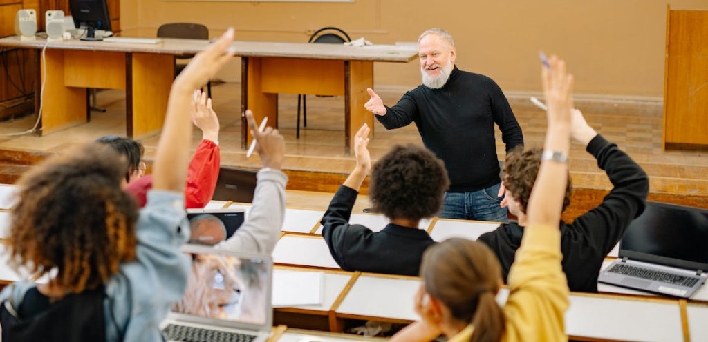 Photo: in a university lecture hall, a smiling professor (a white man of about 40) holds his hand out to the class, as the students raise their hands.